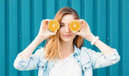 Carefree young beautiful girl using two halfs of oranges instead of glasses over her eyes over blue background.