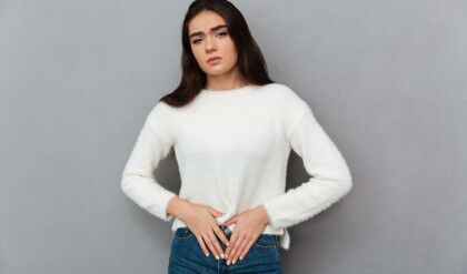 Close-up photo of depressed young woman with abdominal pain, looking at camera, isolated over gray background