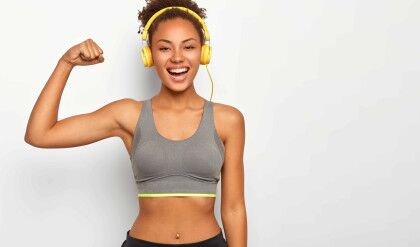 Horizontal view of dark skinned woman in good mood, raises arm with muscles, has strong body, dressed in gym outfit, listens audio via modern headphones, poses indoor. Fitness and music concept