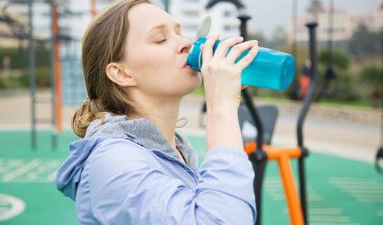 Tired fit girl feeling thirsty during physical exercises. Young woman in sports hoodie drinking water from blue flask or shaker. Fitness or thirst concept