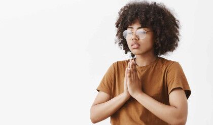 Waist-up shot of focused relaxed and calm attractive young dark-skinned female in glasses with afro hairstyle standing half-turned to left with closed eyes namaste gesture or hands in pray meditating.