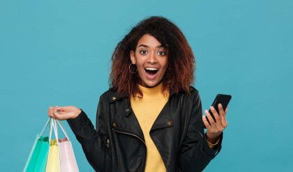 Picture of excited pretty young african woman standing isolated over blue background. Looking camera holding shopping bags and mobile phone.